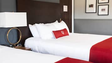 A hotel room with two beds, a bedside table with a lamp, a clock, and framed pictures on the wall.