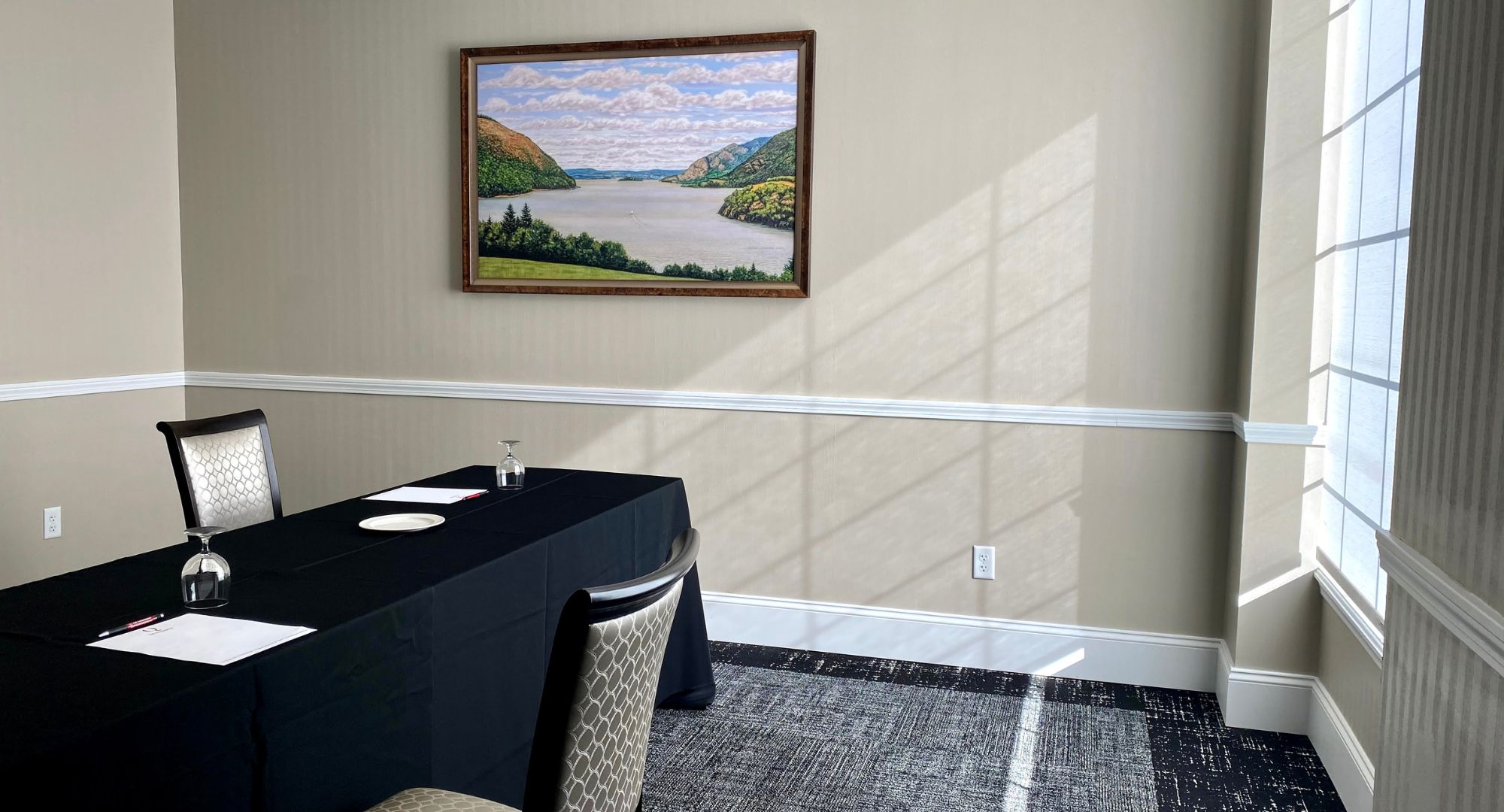 Une salle de conférence avec une table préparée pour une réunion, un tableau représentant un paysage encadré au mur et une fenêtre laissant entrer la lumière naturelle.