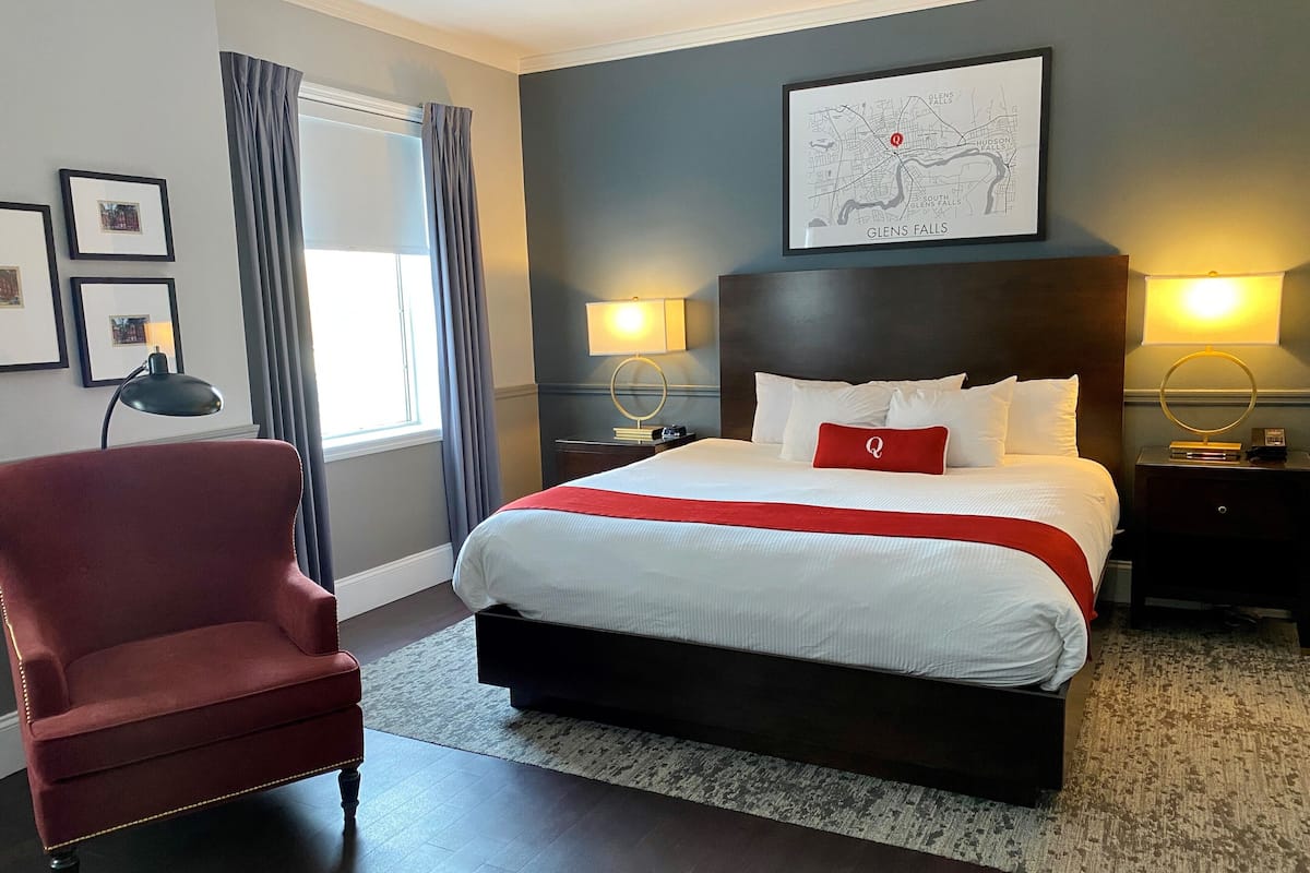 A hotel room with a large bed, a red armchair, two bedside tables with lamps, a map on the wall, and a window with curtains.