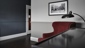 A room with a dark wooden floor, a red chair, a black desk, and a framed black and white photograph on the wall. - The Queensbury Hotel (Glens Falls)