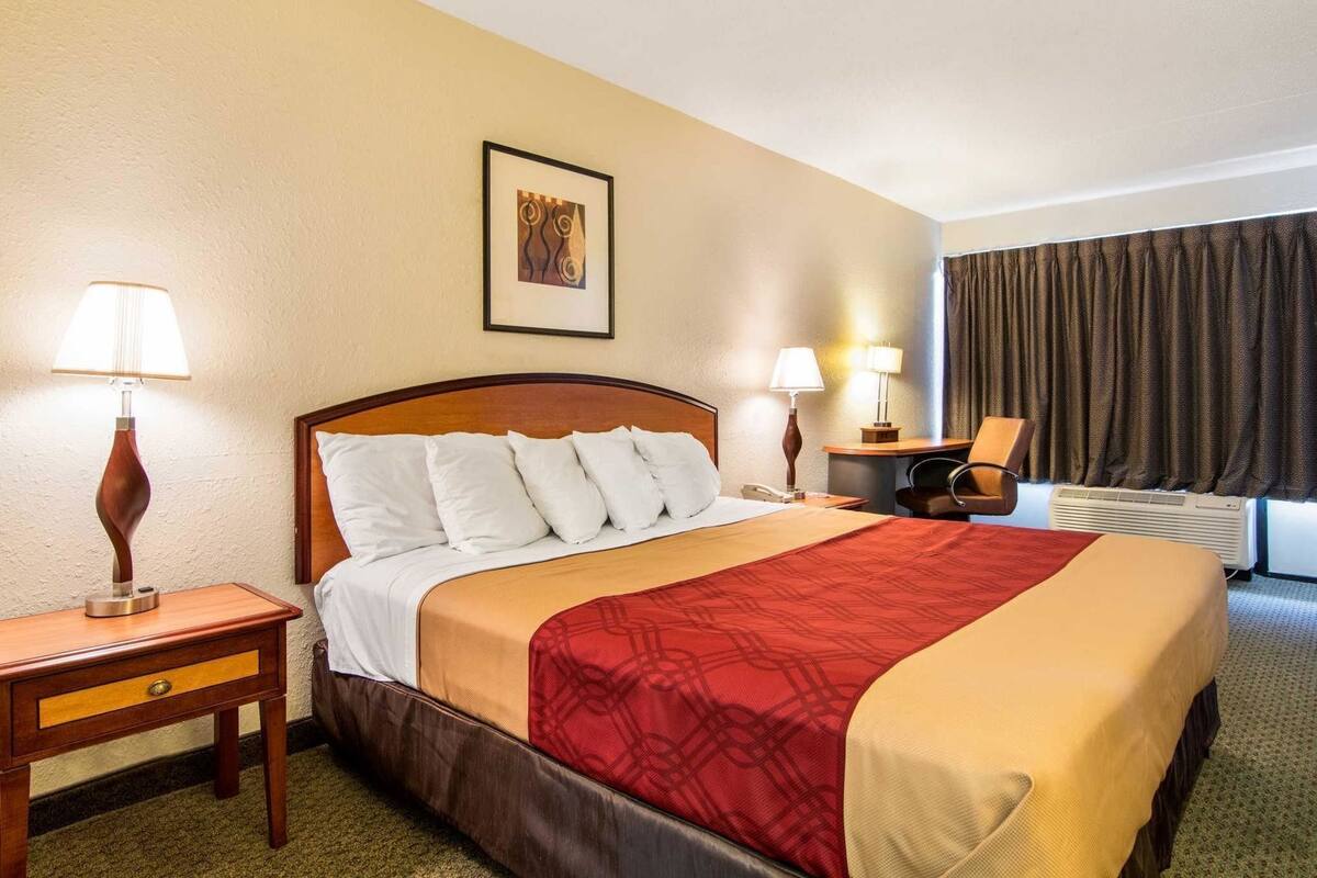 Ein Hotelzimmer mit einem Bett, zwei Nachttischen, einem Schreibtisch mit Stuhl, einem Fenster mit Vorhängen und einem Wandbild.