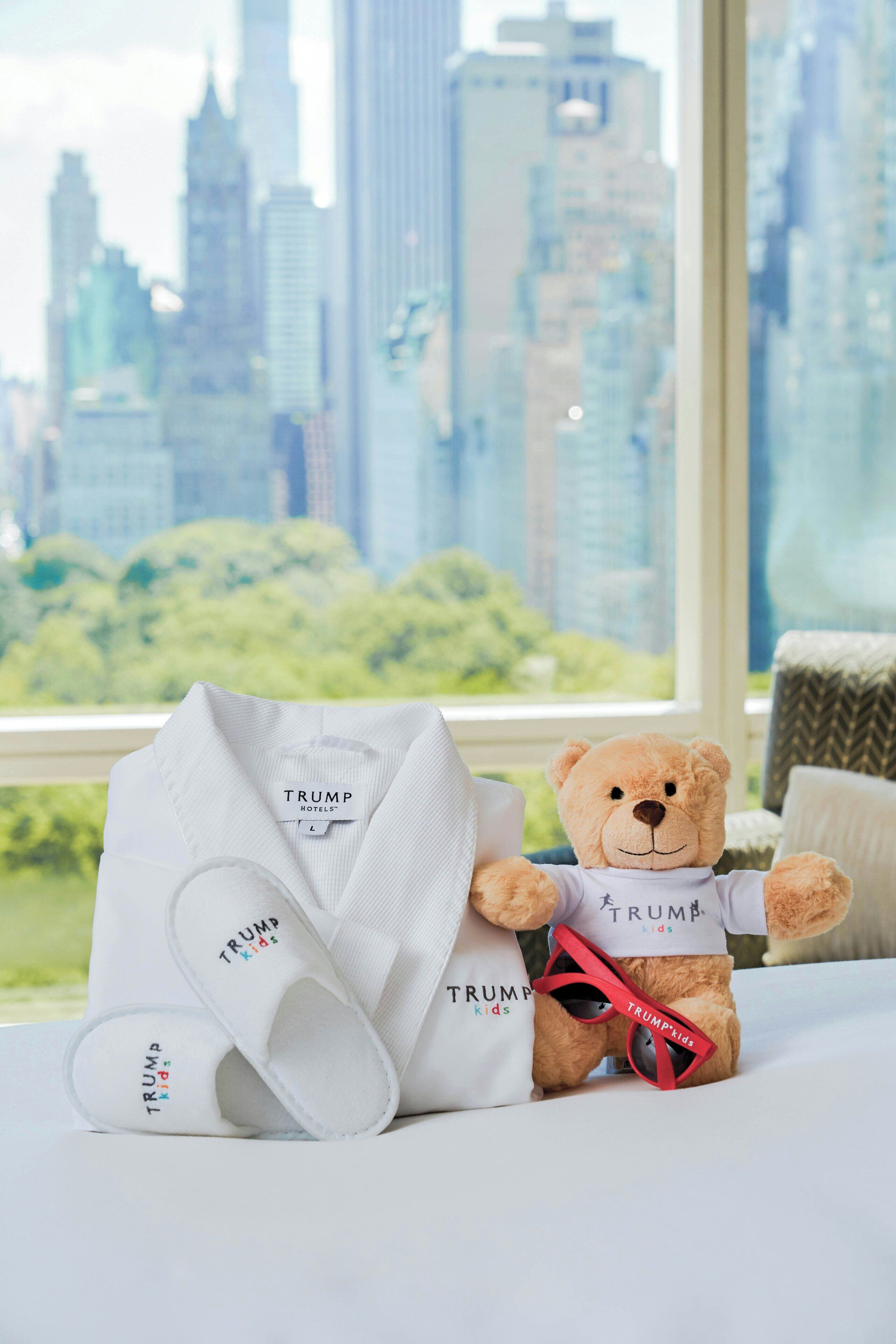 A teddy bear wearing a white shirt with 'TRUMP' printed on it, placed on a bed with a cityscape view through a window.