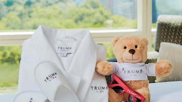 A teddy bear wearing a white shirt with 'TRUMP' printed on it, placed on a bed with a cityscape view through a window.