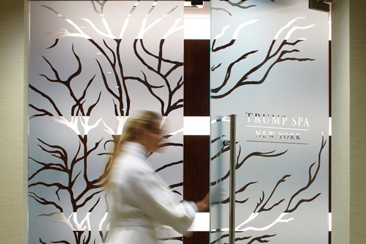 Een vrouw in een witte badjas loopt langs een deur met een boommotief en de tekst 'TRUMP SPA NEW YORK'.