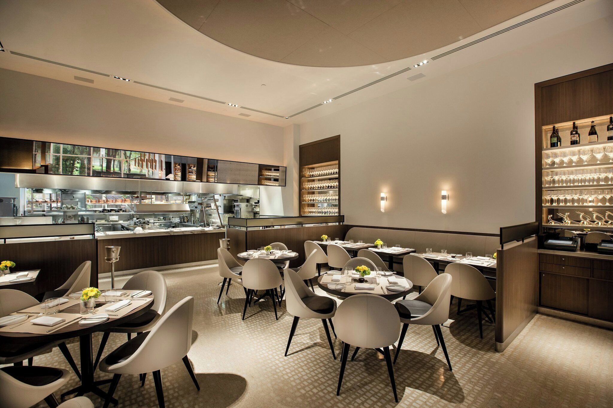 A modern restaurant interior with round tables, white chairs, and a bar area with wine glasses.