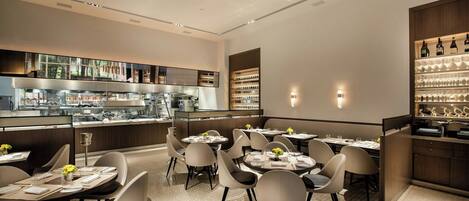 A modern restaurant interior with round tables, white chairs, and a bar area with wine glasses.