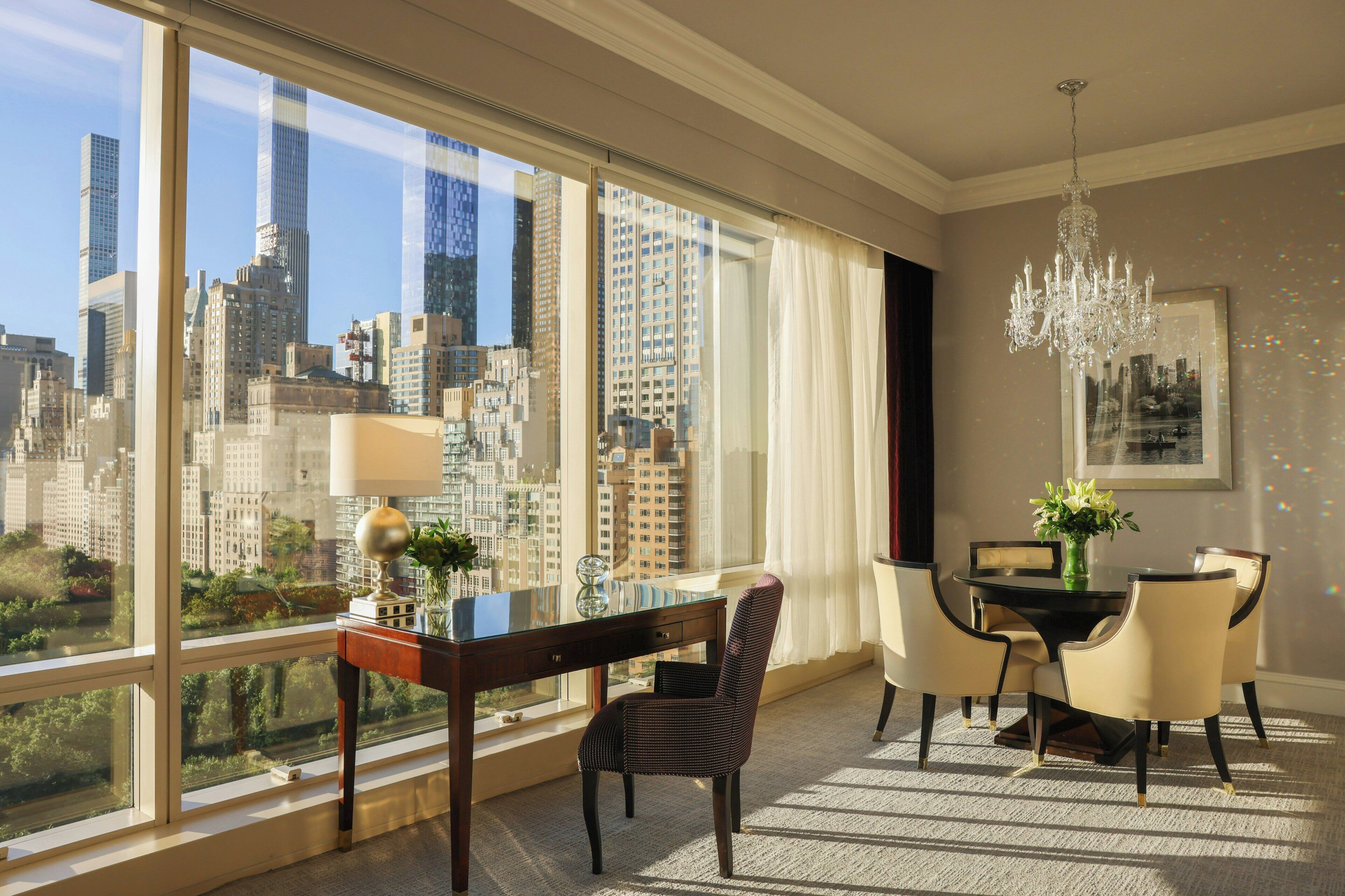 A hotel room with a large window offering a city view, a dining area with a chandelier, and a table with chairs.
