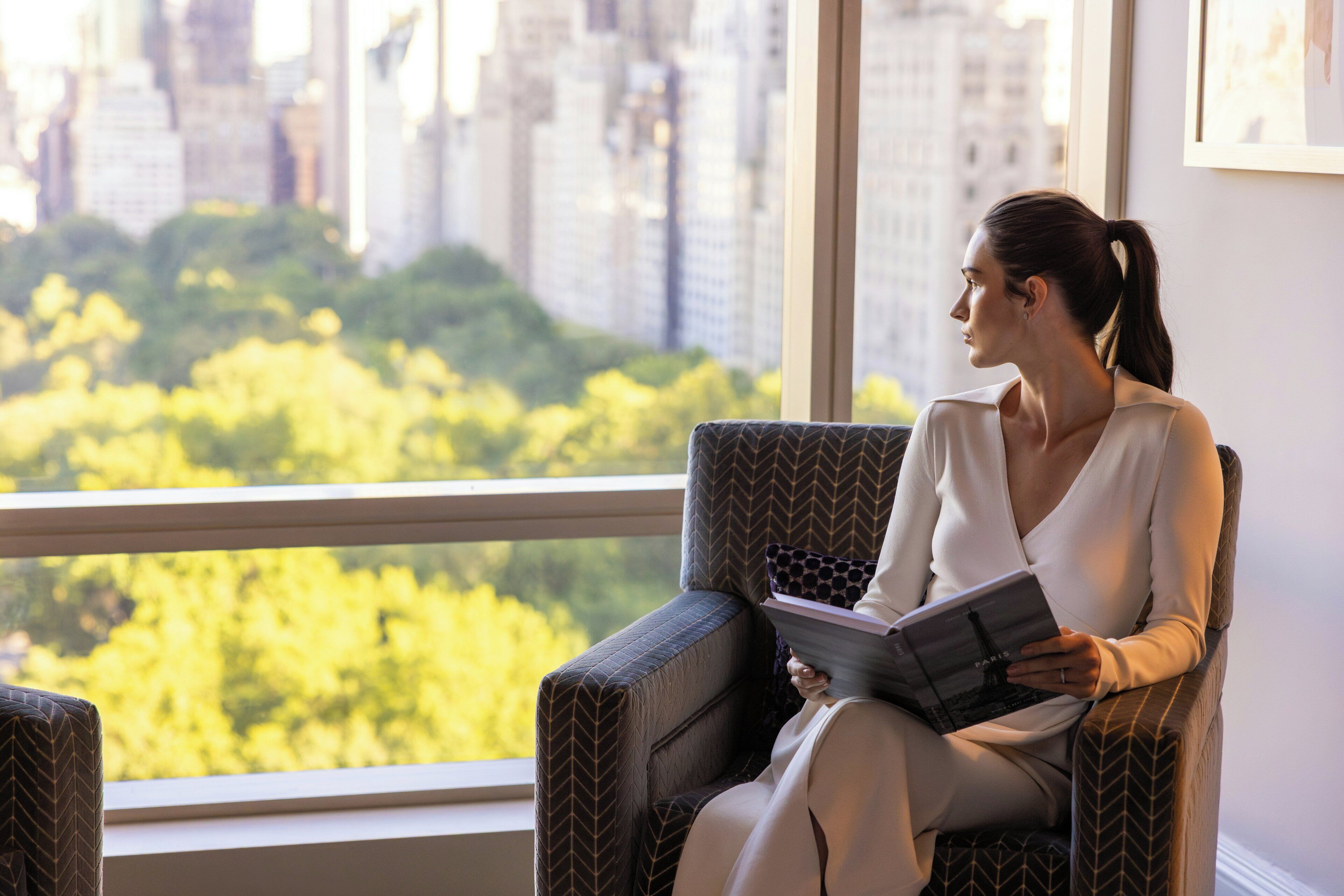 A person seated in a chair with a book, overlooking a cityscape and greenery through a window.