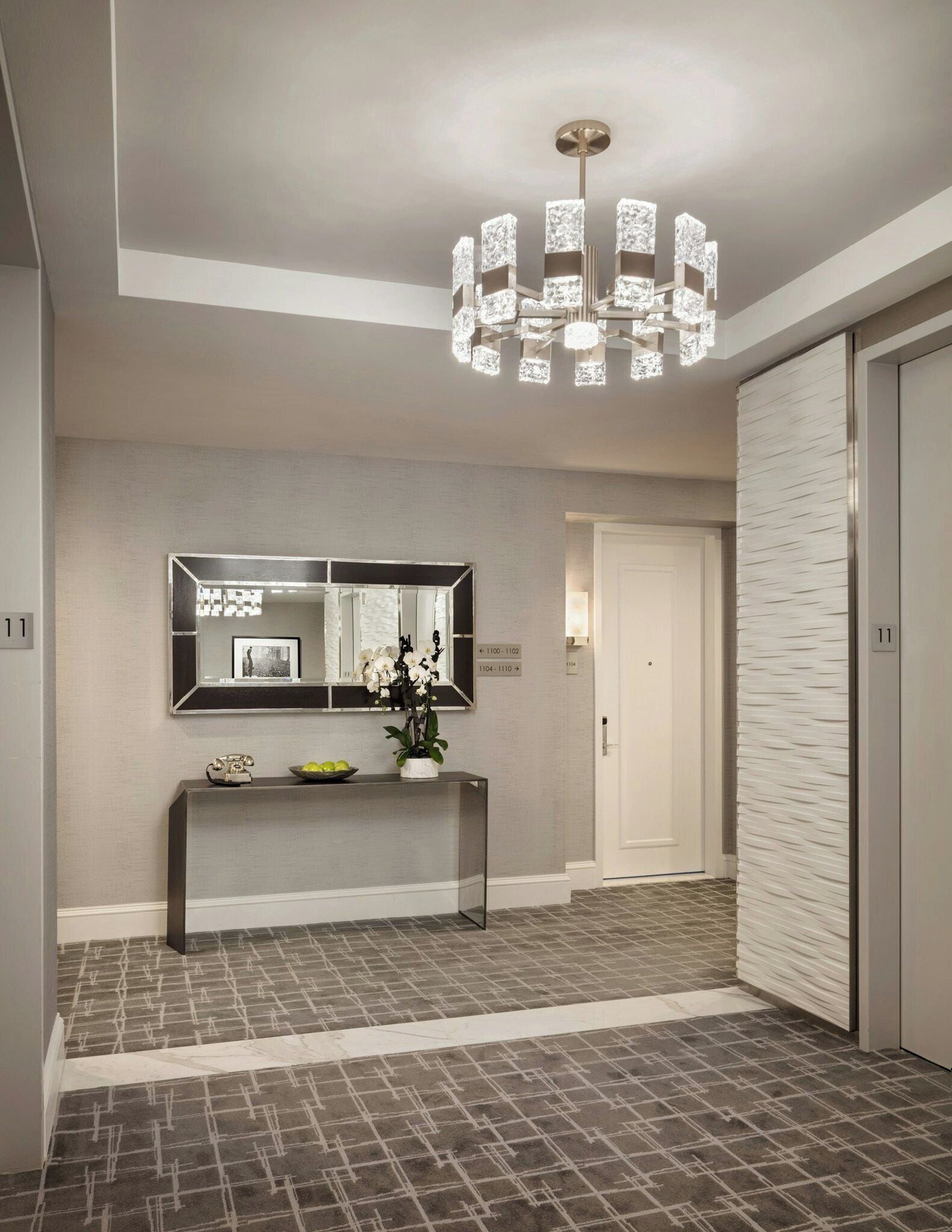A modern hallway with a large mirror, a console table with decorative items, a patterned floor, and a unique chandelier.