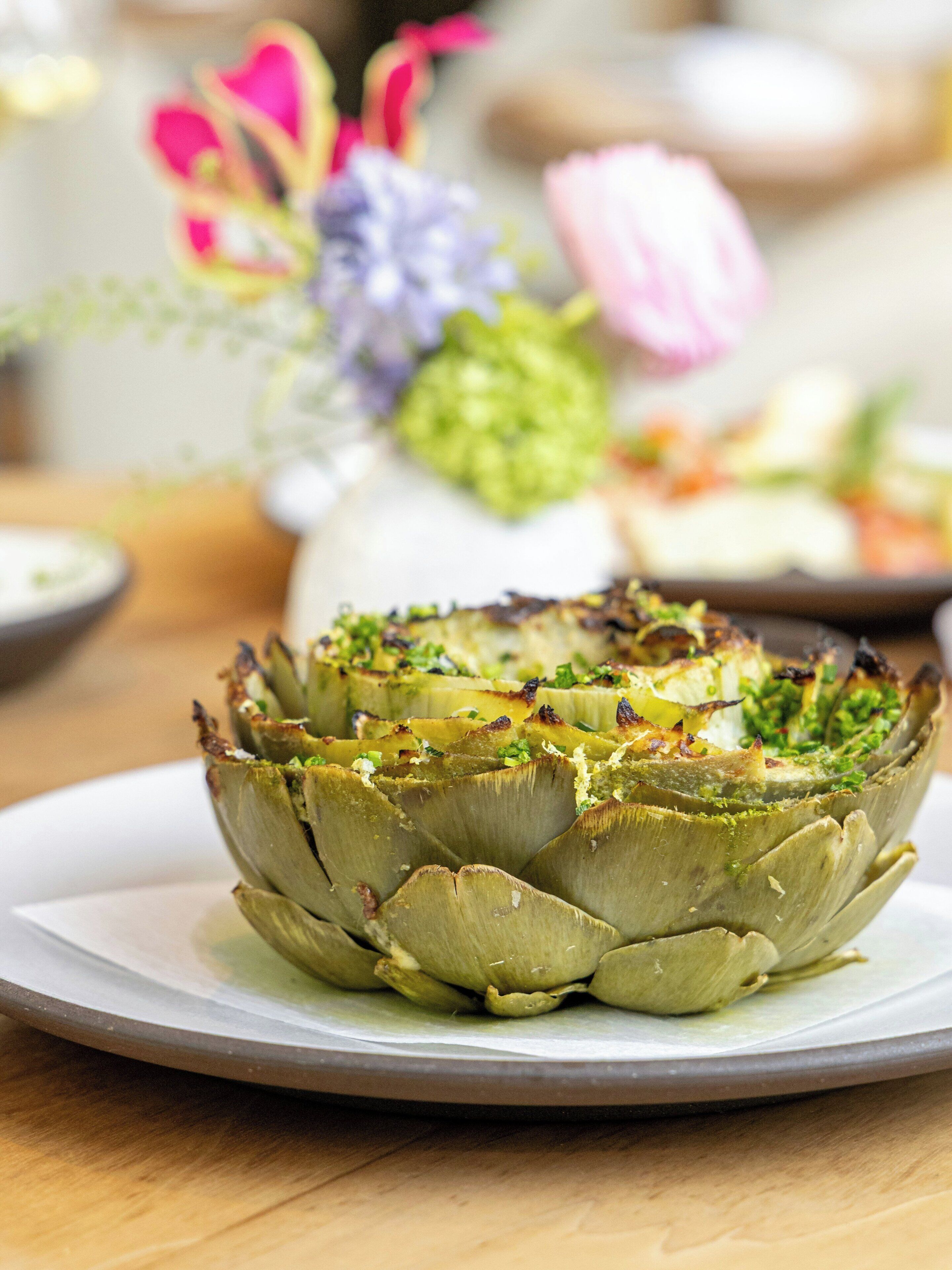 A plate with a roasted artichoke centerpiece, a white plate, and a wooden table surface.