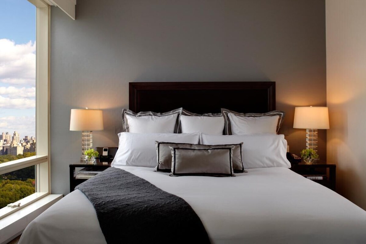 Een hotelkamer met een groot bed, twee bedlampen en uitzicht op een stadsbeeld door een groot raam.