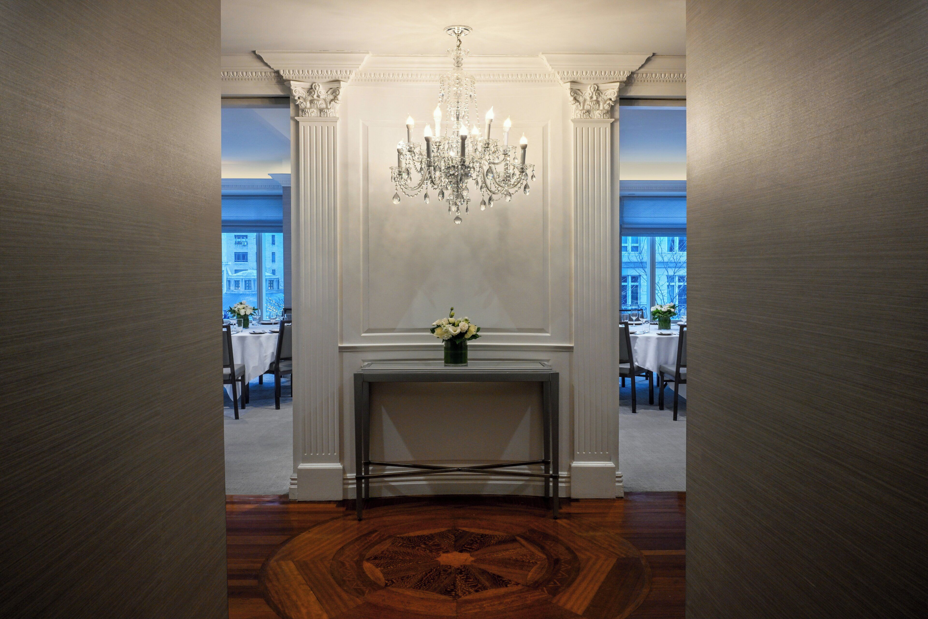 A hallway with a central console table, a chandelier, and two tables set for dining with chairs.