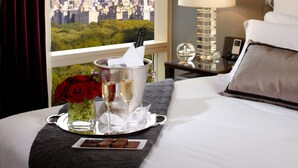Une chambre d’hôtel avec vue sur la ville, un lit, une table de chevet avec une lampe, un plateau avec un vase de roses rouges, des coupes à champagne et une assiette de biscuits.
