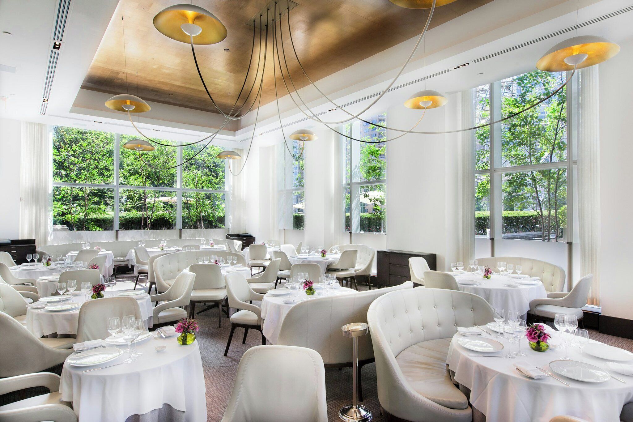 A spacious dining area with multiple tables and chairs, large windows with a view of greenery, and modern pendant lighting.