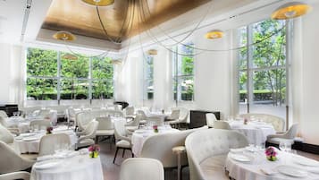 A spacious dining area with multiple tables and chairs, large windows with a view of greenery, and modern pendant lighting.