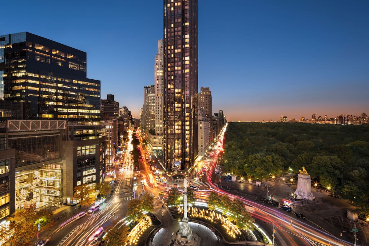 A nighttime cityscape with a prominent skyscraper, busy streets, and a park with a fountain.