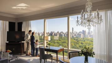 A spacious hotel room with a large window offering a city view, a chandelier, and a dining area with a table and chairs.