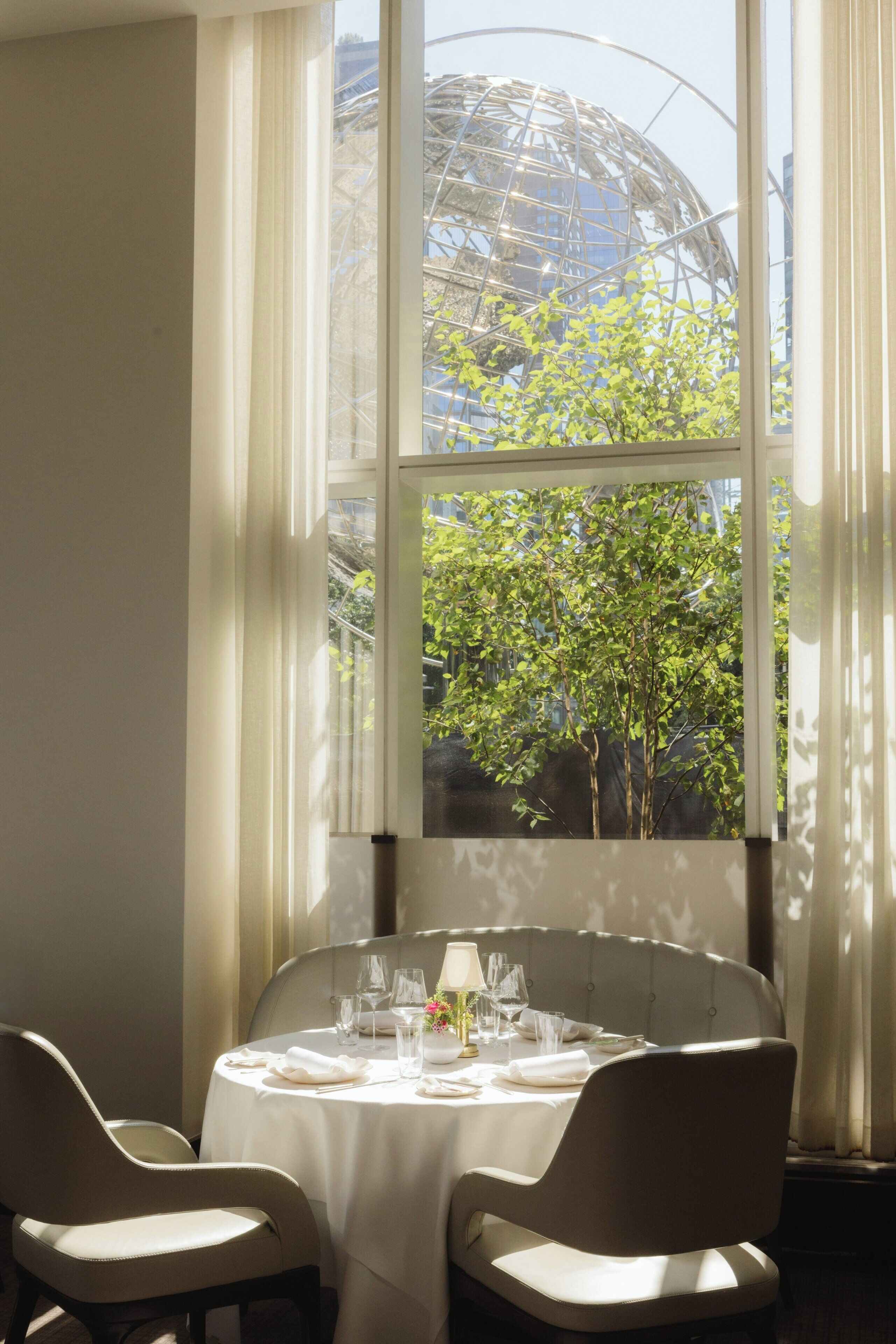 Un coin repas avec une table ronde dressée pour quatre personnes, une fenêtre donnant sur un bâtiment et sur de la verdure, et des rideaux légers.