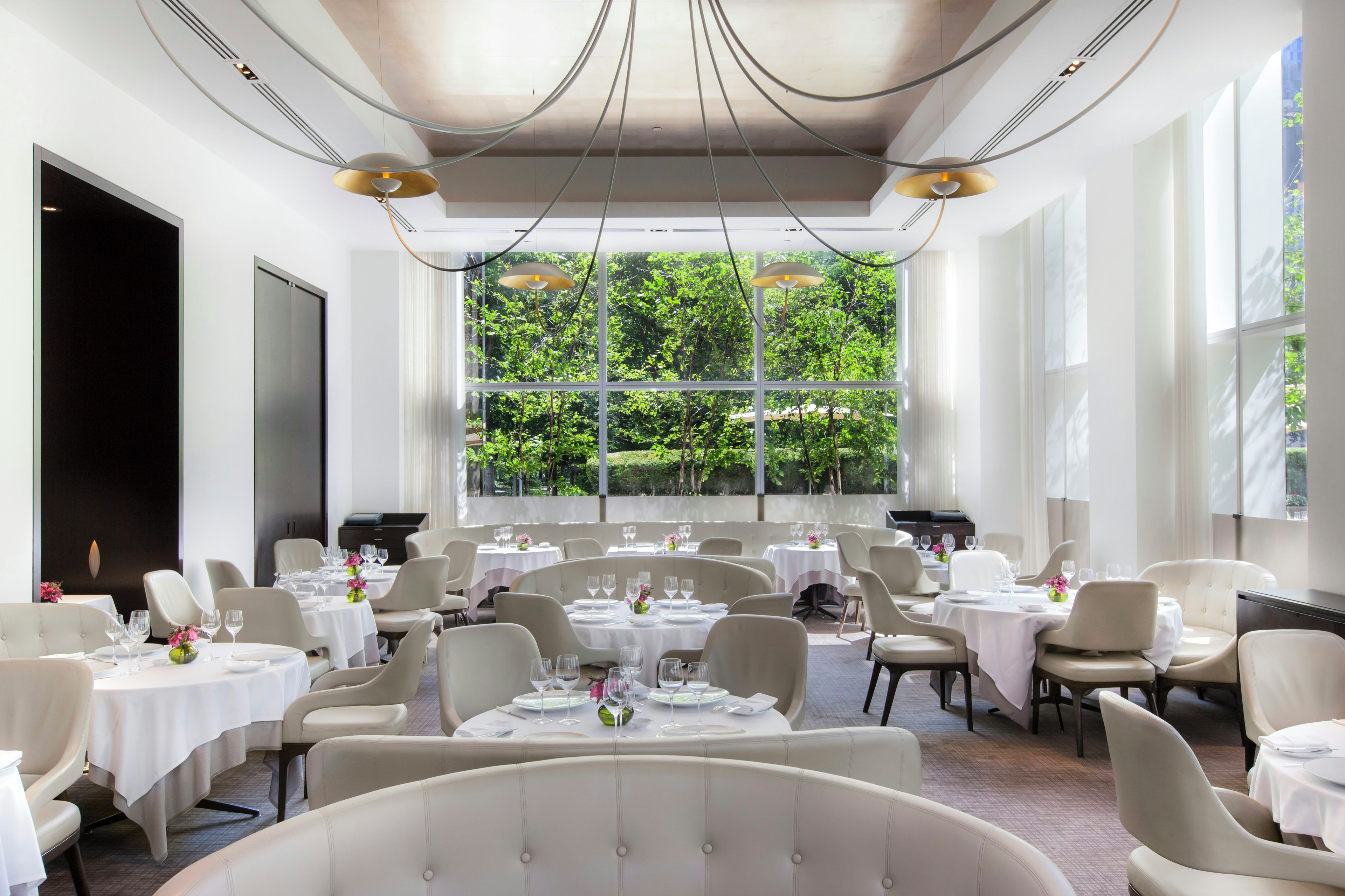 Un restaurante con varias mesas y sillas, amplios ventanales con vistas a zonas verdes e iluminación colgante moderna.