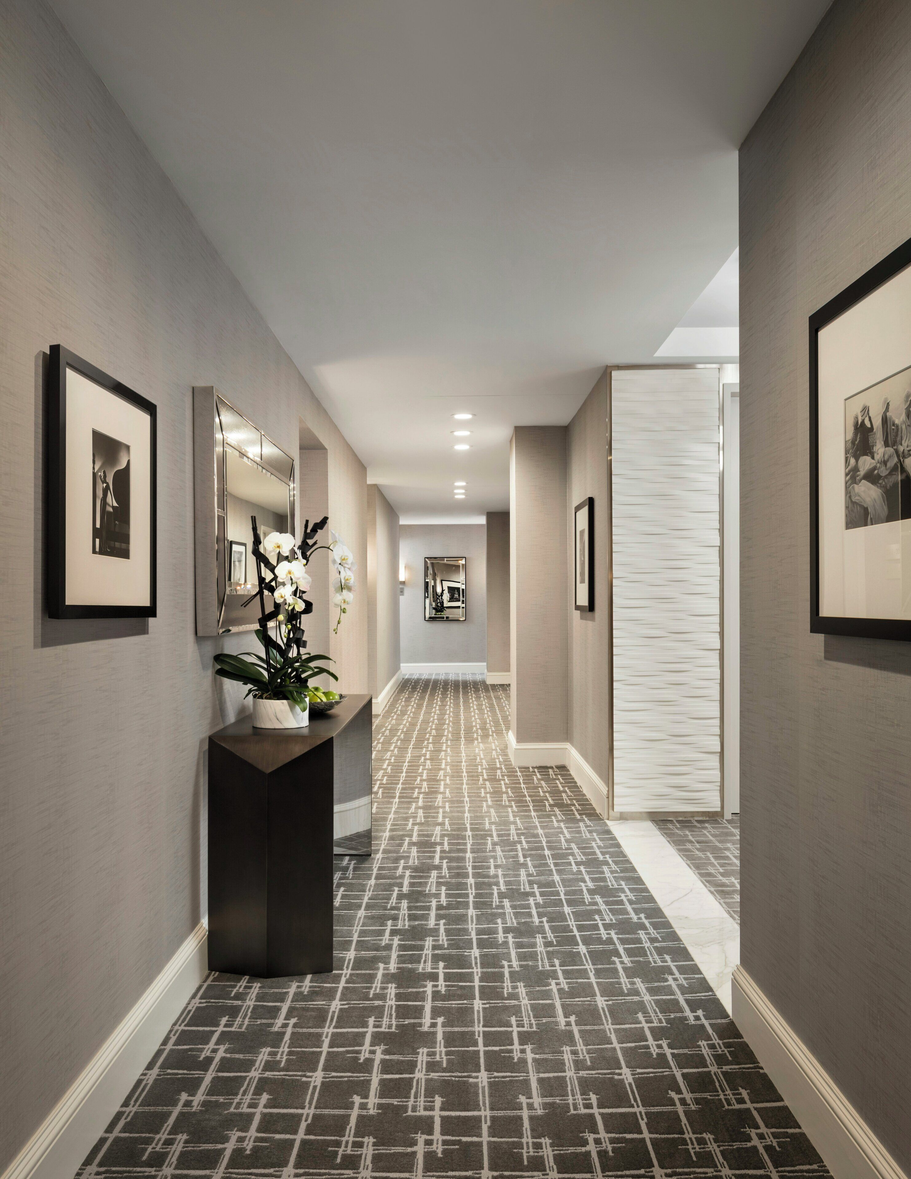 A hallway with patterned carpet, framed artwork on the walls, a potted plant, and a mirror.