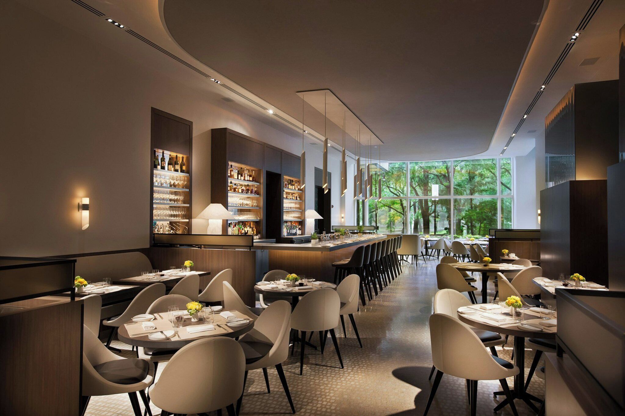 A modern restaurant with a long counter, multiple tables, and a view of trees through large windows.