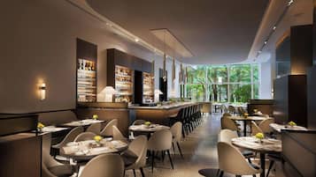 A modern restaurant with a long counter, multiple tables, and a view of trees through large windows.