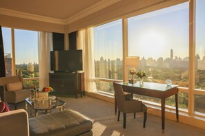 Habitación de hotel moderna con un amplio ventanal que ofrece vistas a la ciudad, un televisor de pantalla plana, una mesa de comedor con un jarrón de flores y una zona de estar acogedora.