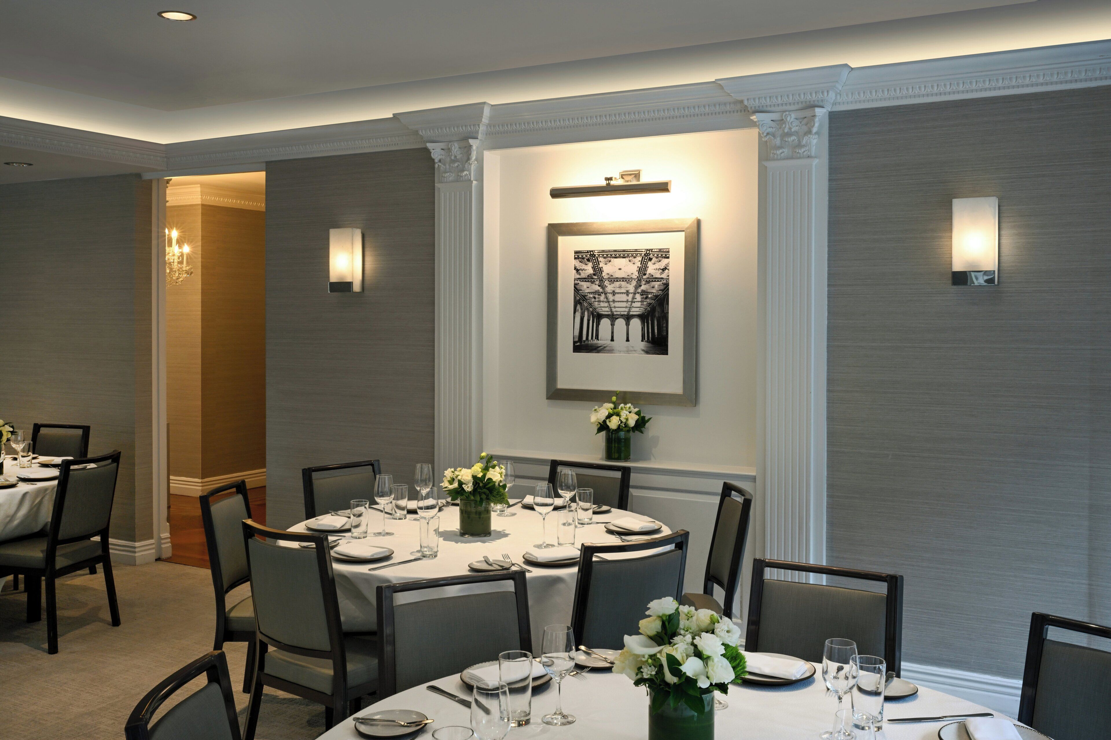 A dining area with a round table set for six, surrounded by chairs, with a framed picture above the table and wall-mounted lighting on either side.