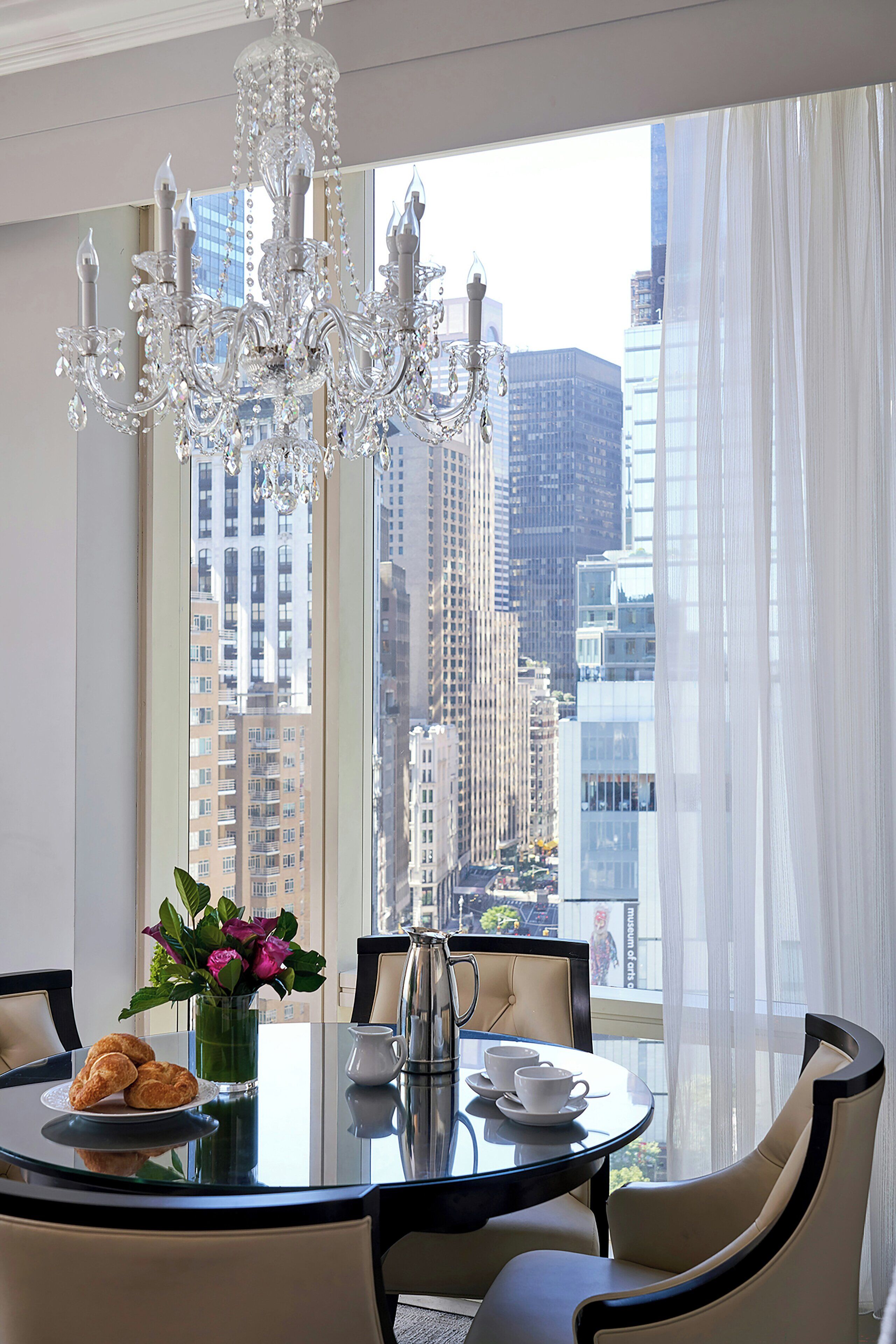 A dining area with a chandelier, a table set for two, and a view of a cityscape through large windows.