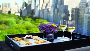 Un plateau avec du fromage, des noix et un bol de fruits rouges, deux verres de vin blanc et un vase de fleurs violettes, le tout placé devant un paysage urbain.