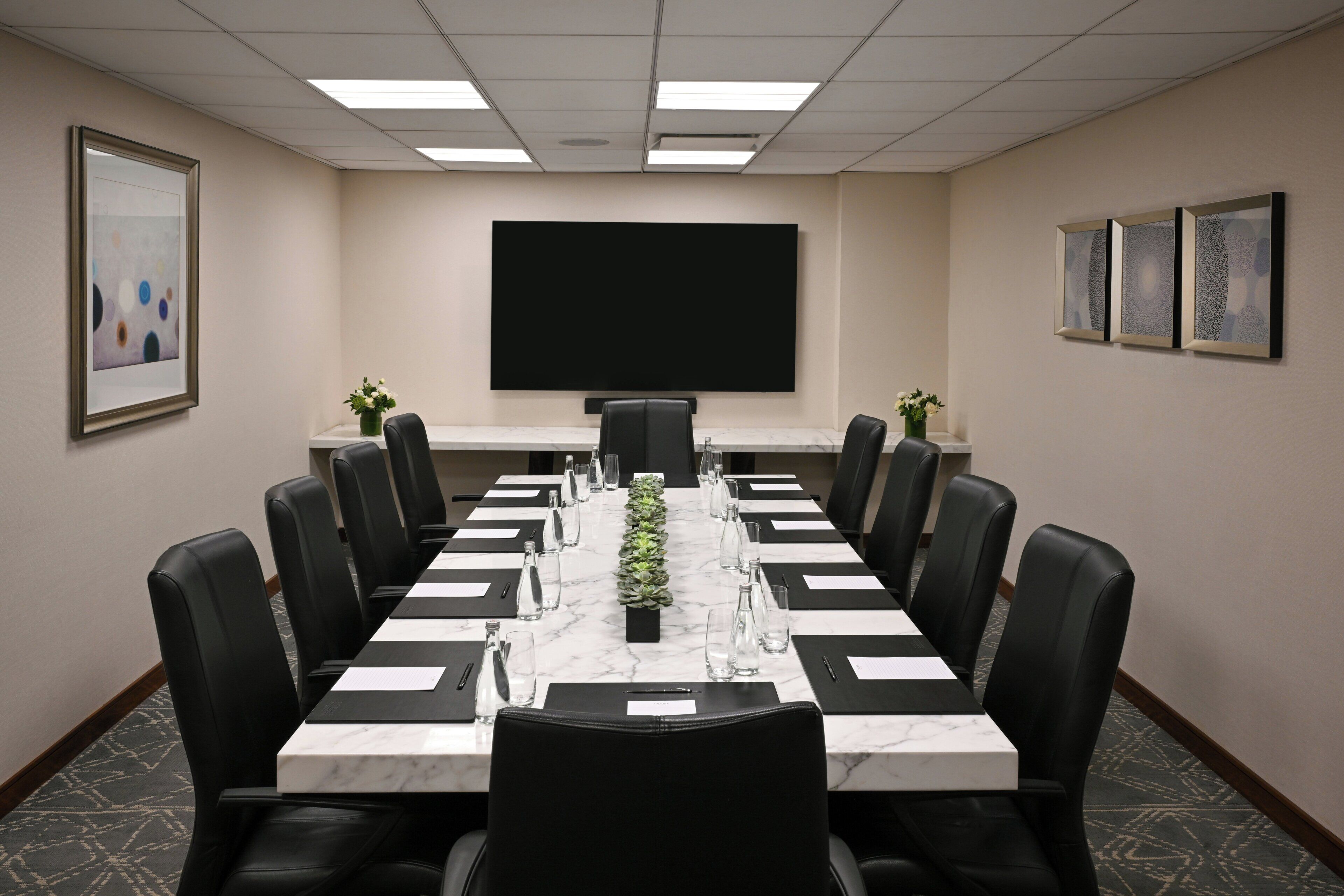A conference room with a long table, black leather chairs, a projector screen, and framed artwork on the walls.