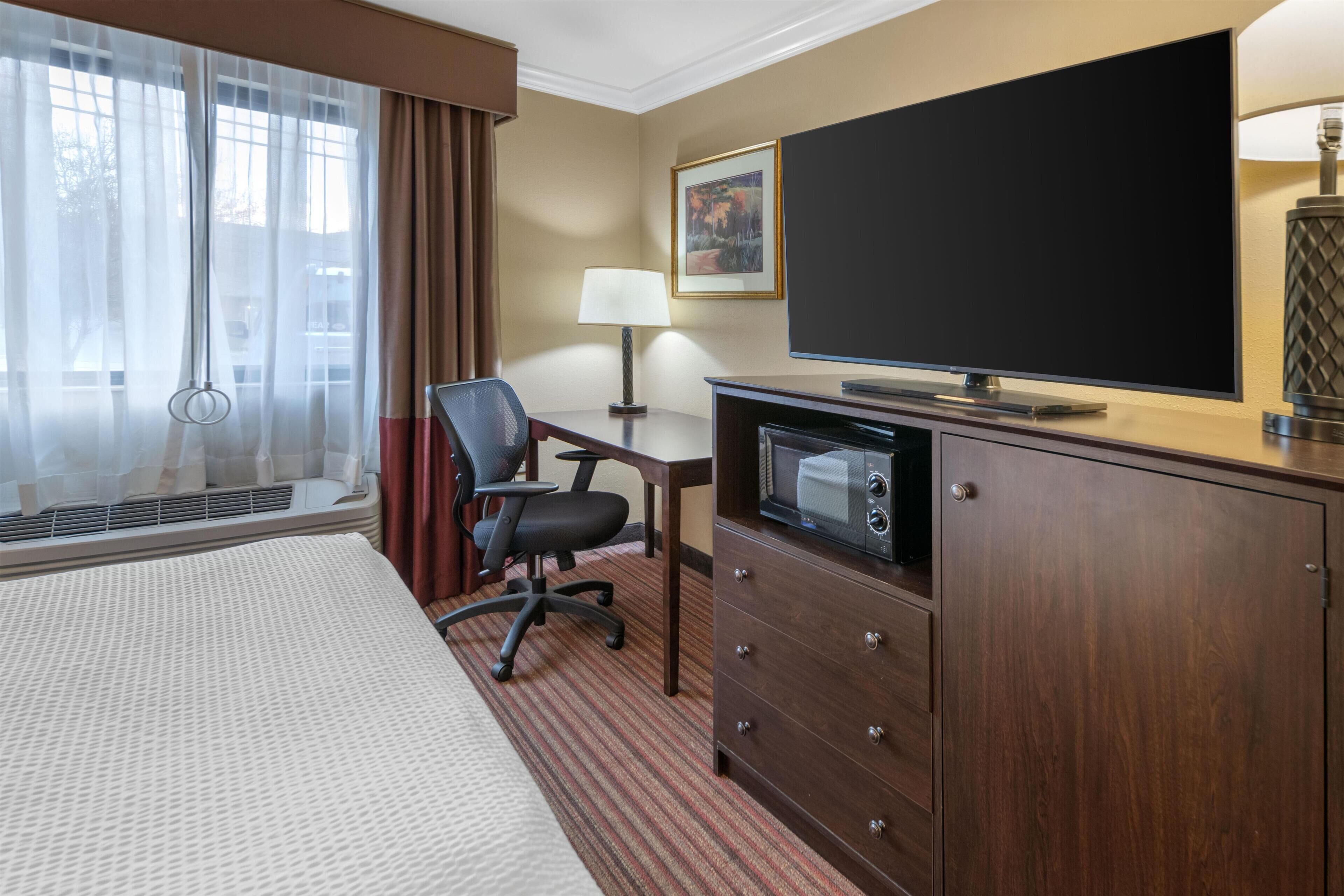 Una habitación de hotel con cama, escritorio, televisión, silla, lámpara y un cuadro en la pared.