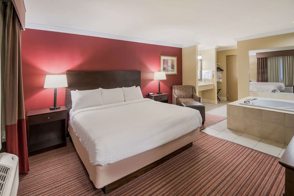 A hotel room with a large bed, two bedside tables with lamps, a chair, a bathroom with a bathtub, and a mirror.
