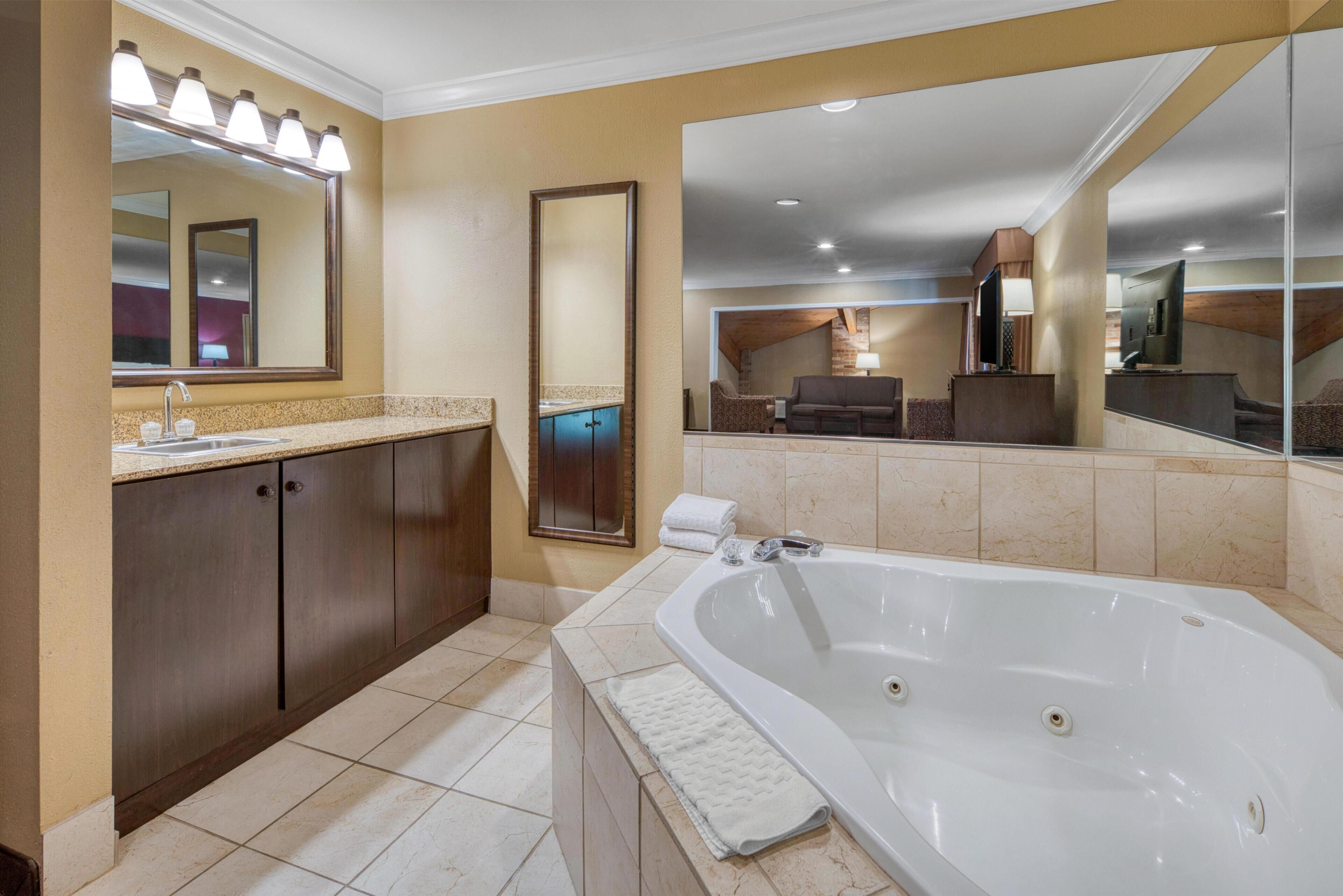 A modern bathroom with a large bathtub, a vanity with a mirror, and a seating area visible through the mirror.