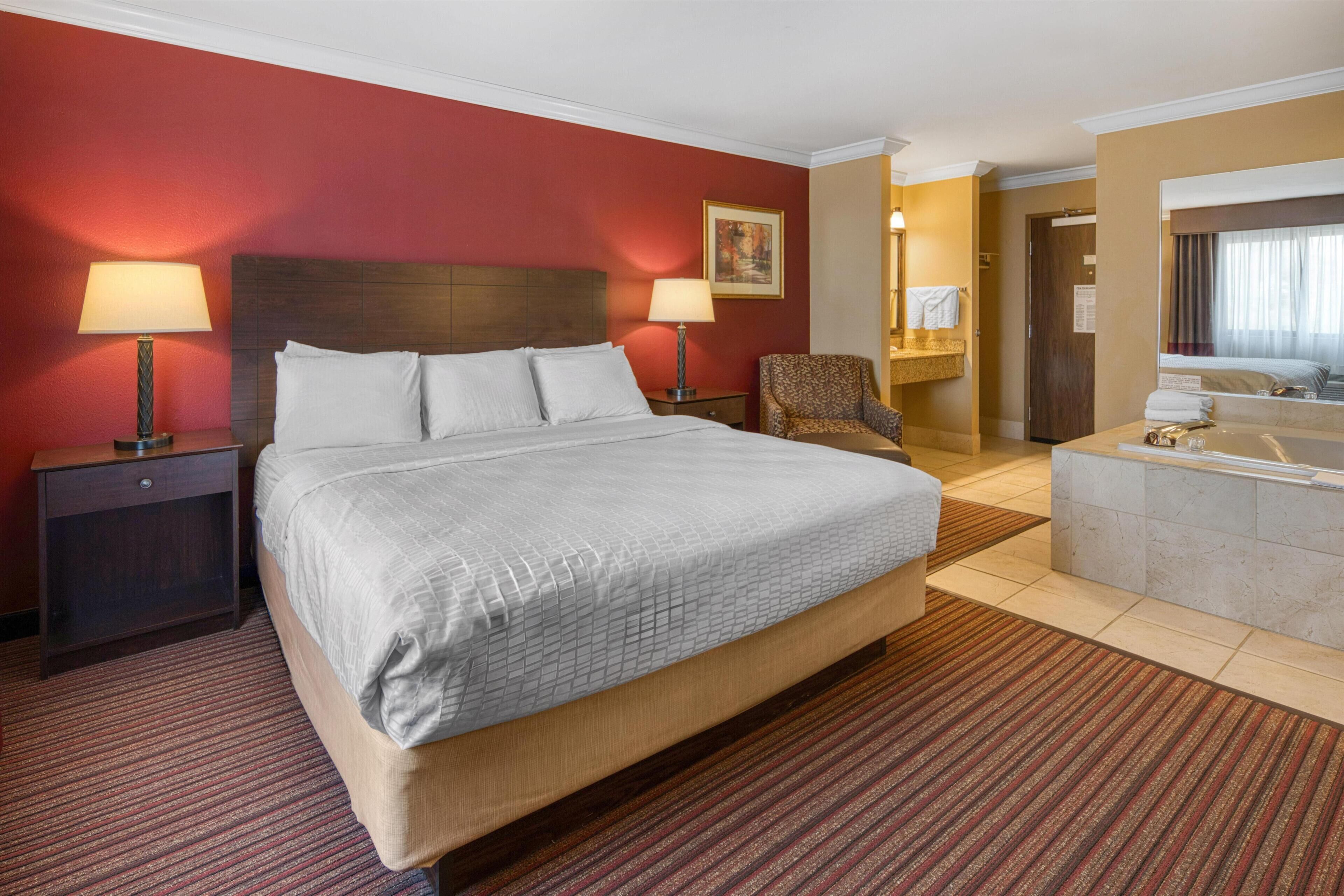 A hotel room with a large bed, two bedside tables with lamps, a chair, a bathroom with a bathtub, and a door leading to another room.