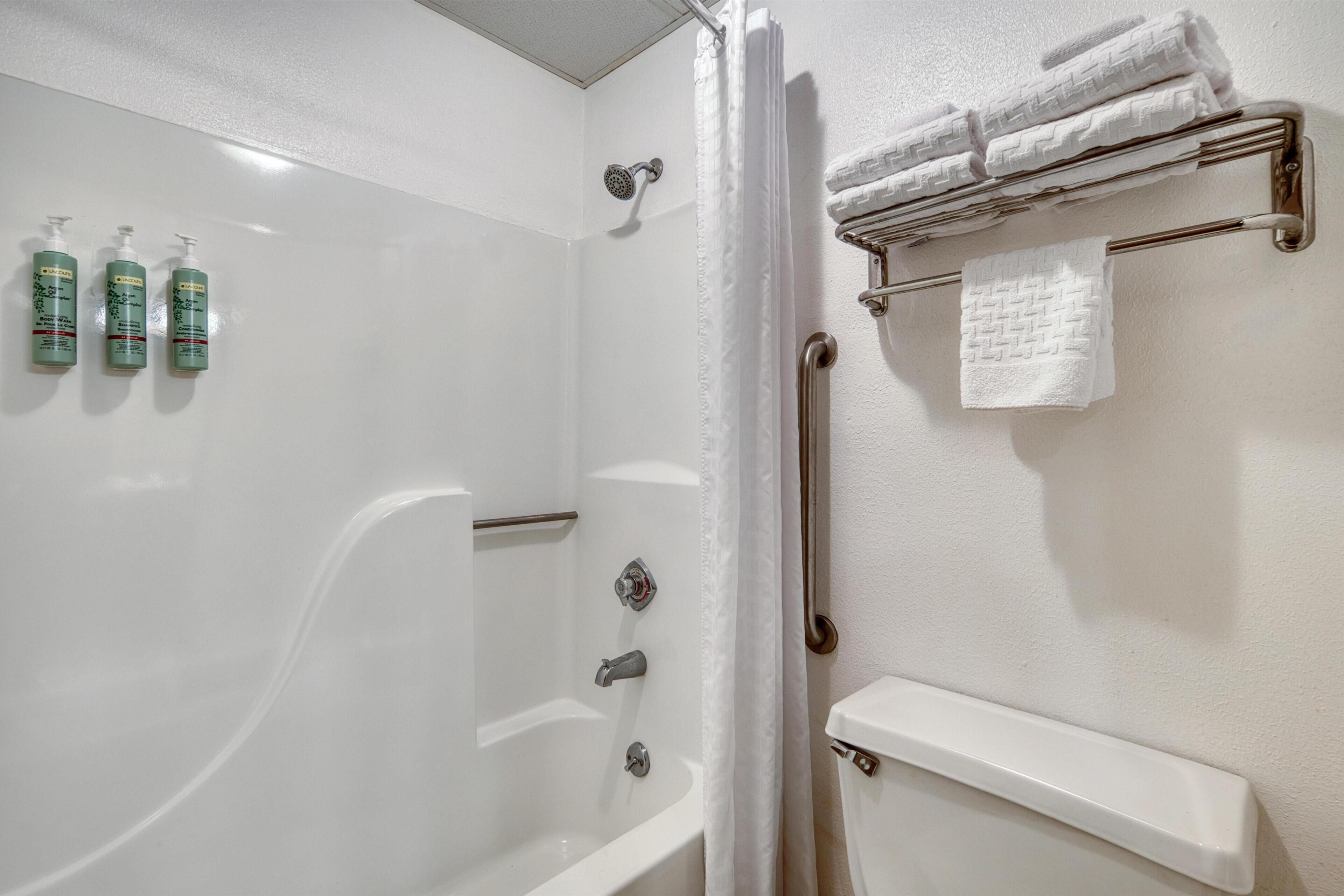 A bathroom with a bathtub, shower, and handrails. There are three bottles of shampoo on the wall.