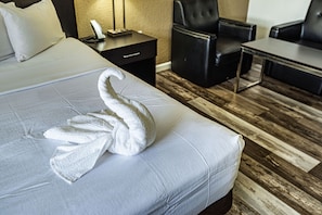 A hotel room with a neatly made bed, a black leather armchair, a small table, and a nightstand with a lamp.