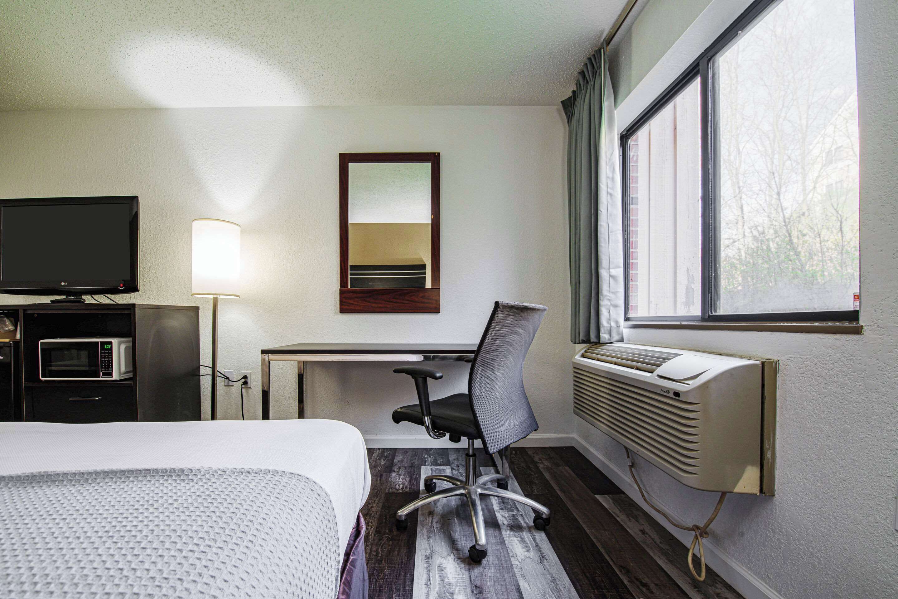 Une chambre d’hôtel comprenant un lit, un bureau avec une chaise, une télévision, une lampe et une fenêtre avec des rideaux.