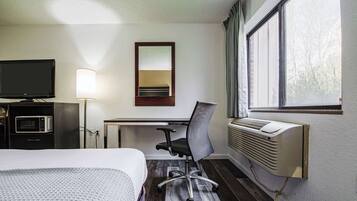 Une chambre d’hôtel comprenant un lit, un bureau avec une chaise, une télévision, une lampe et une fenêtre avec des rideaux.