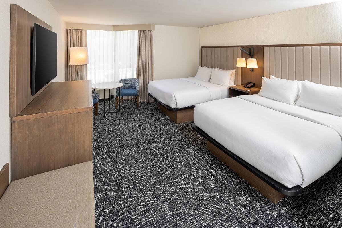 A hotel room with two beds, a flat-screen TV, a desk with a chair, and a window with curtains.