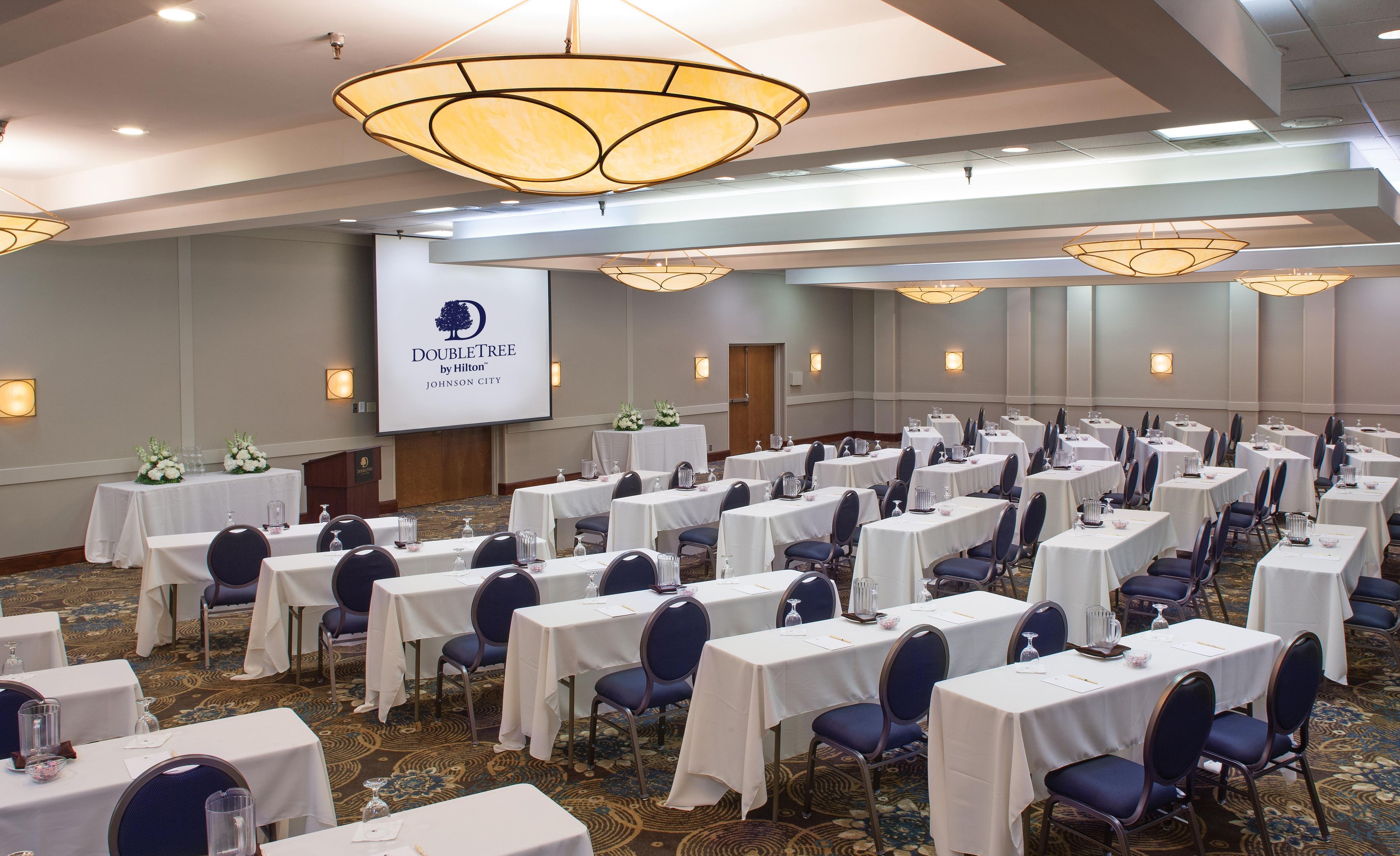 A conference room with multiple tables, chairs, and a large screen displaying the DoubleTree by Hilton logo.