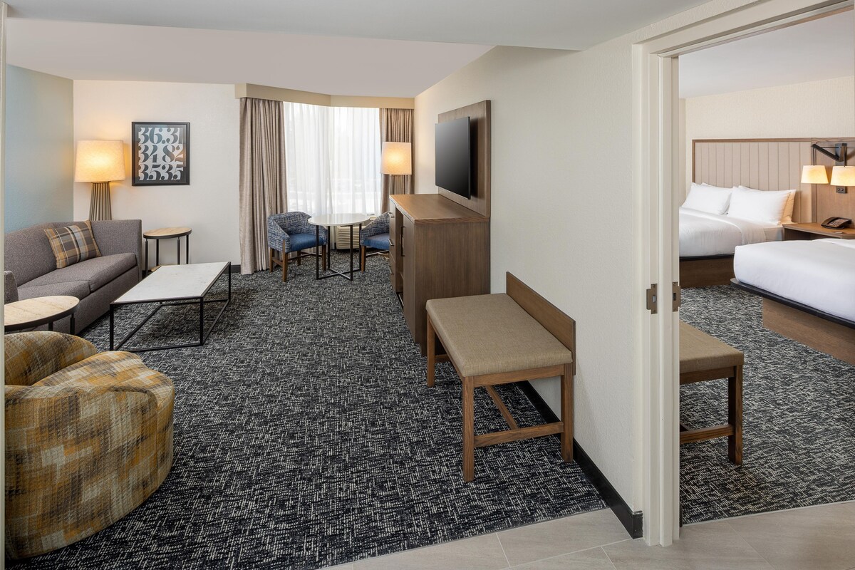 A hotel room with a sofa, two armchairs, a small table, a bench, a TV, a desk, and a bed.