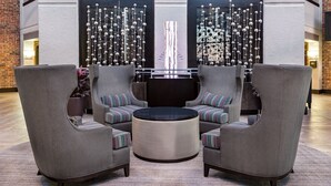 Un área de recepción con una disposición de tres sillones grises y una mesa redonda, una pared decorativa con elementos colgantes y una pared de ladrillo en un lateral.