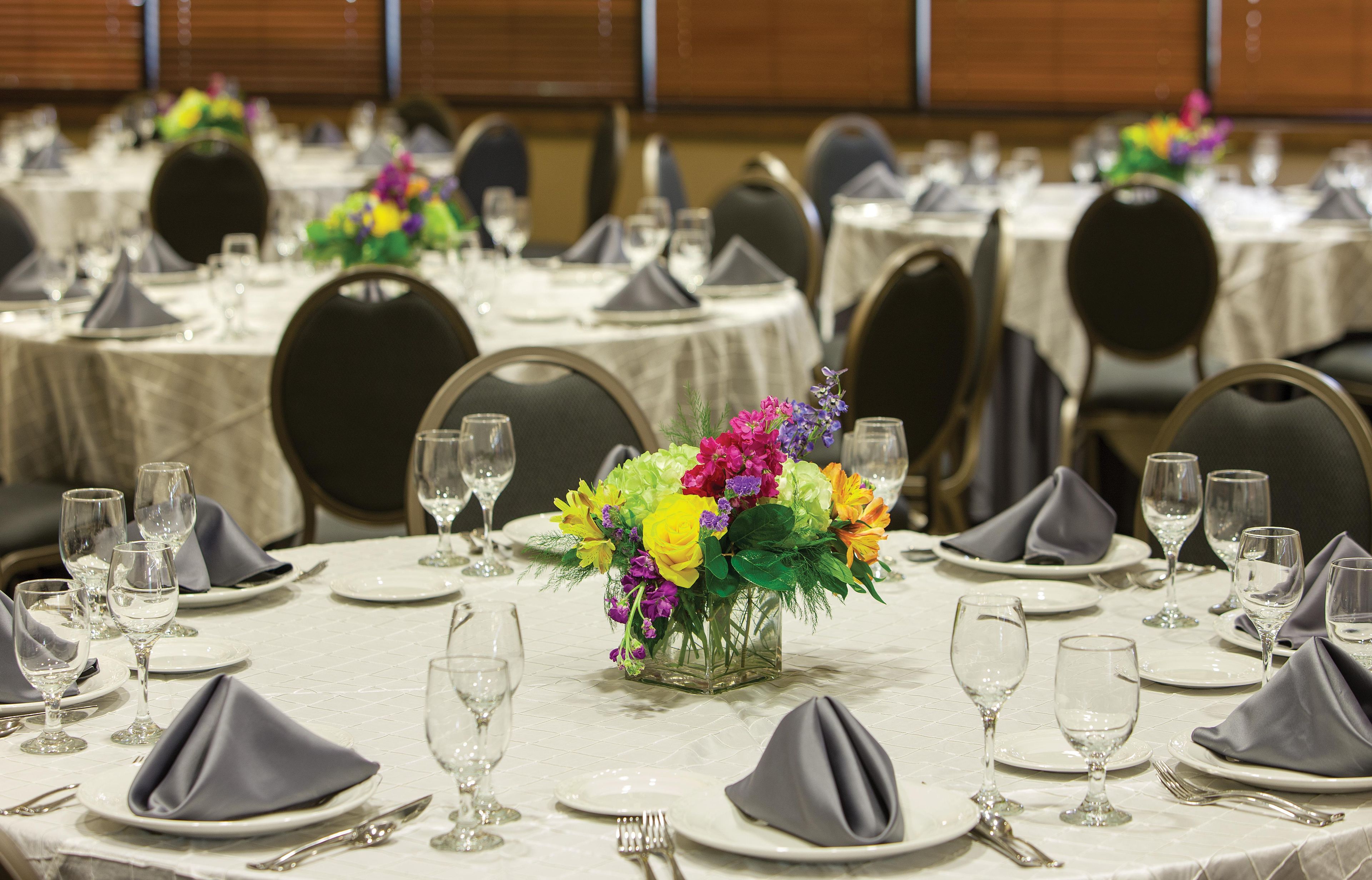 A banquet hall with round tables set for dining, each with a vase of flowers, plates, cutlery, and napkins.