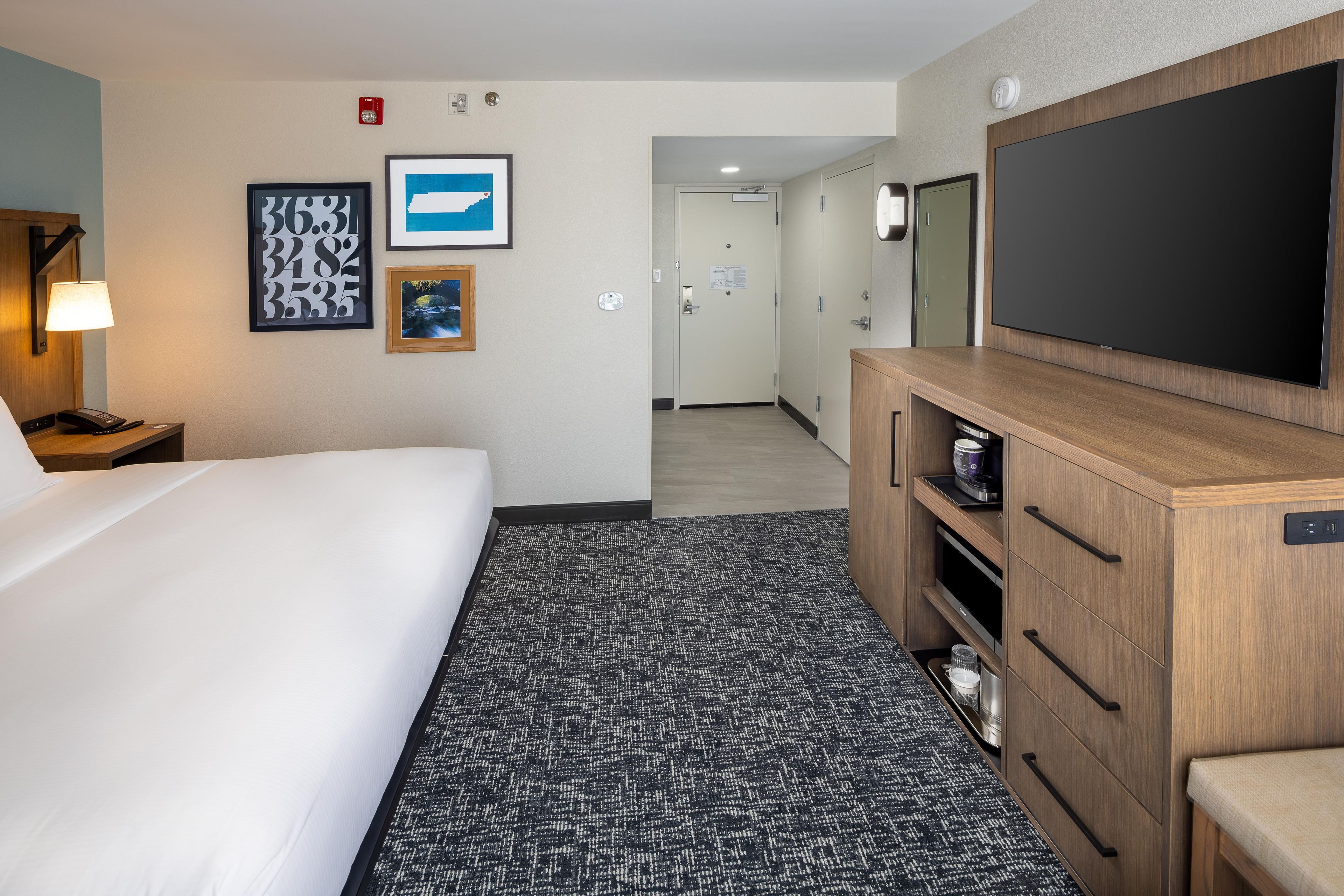 Ein Hotelzimmer mit Bett, Schreibtisch mit Lampe, Fernseher, Kommode und Teppichboden.