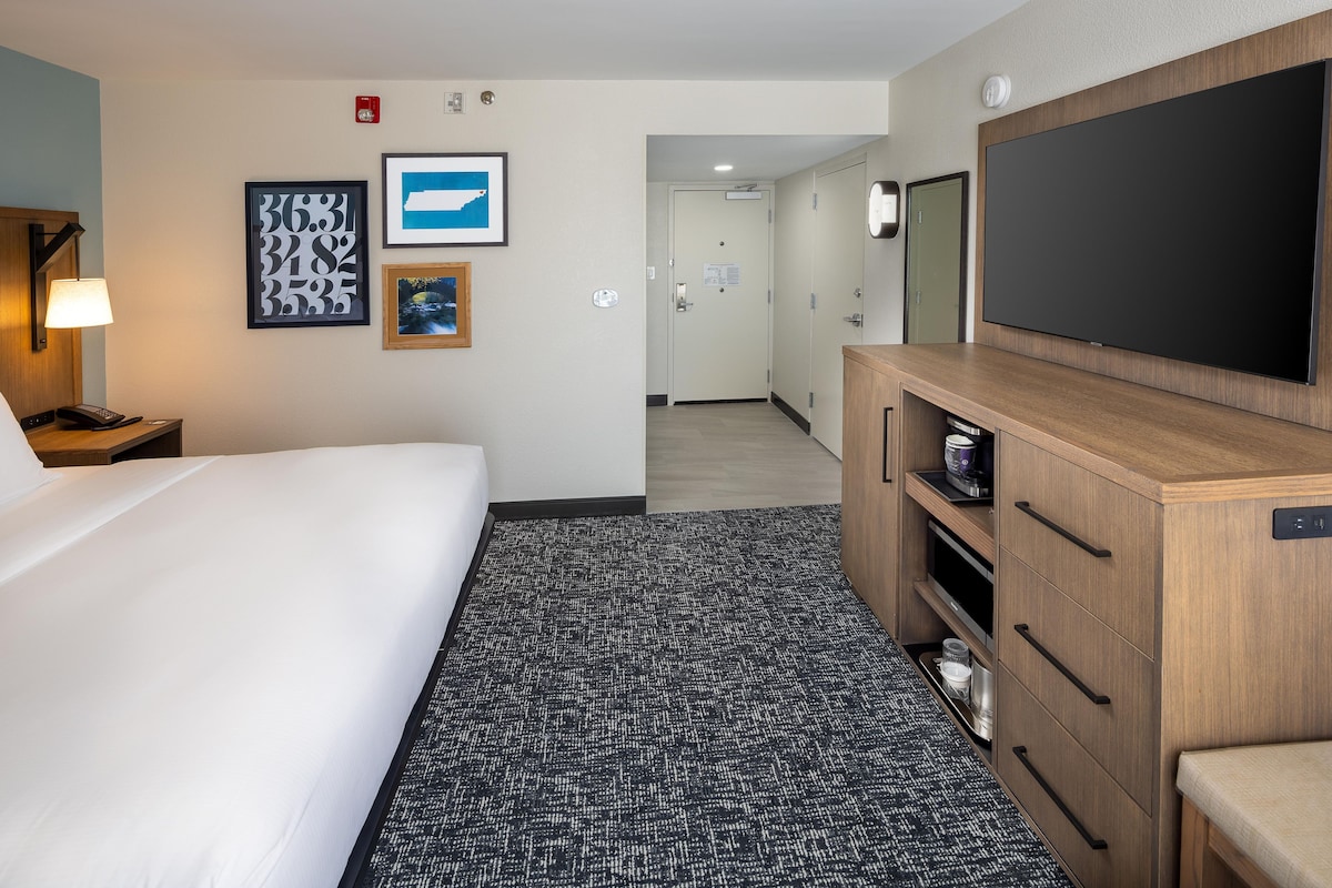 A hotel room with a bed, a desk with a lamp, a TV, a dresser, and a carpeted floor.