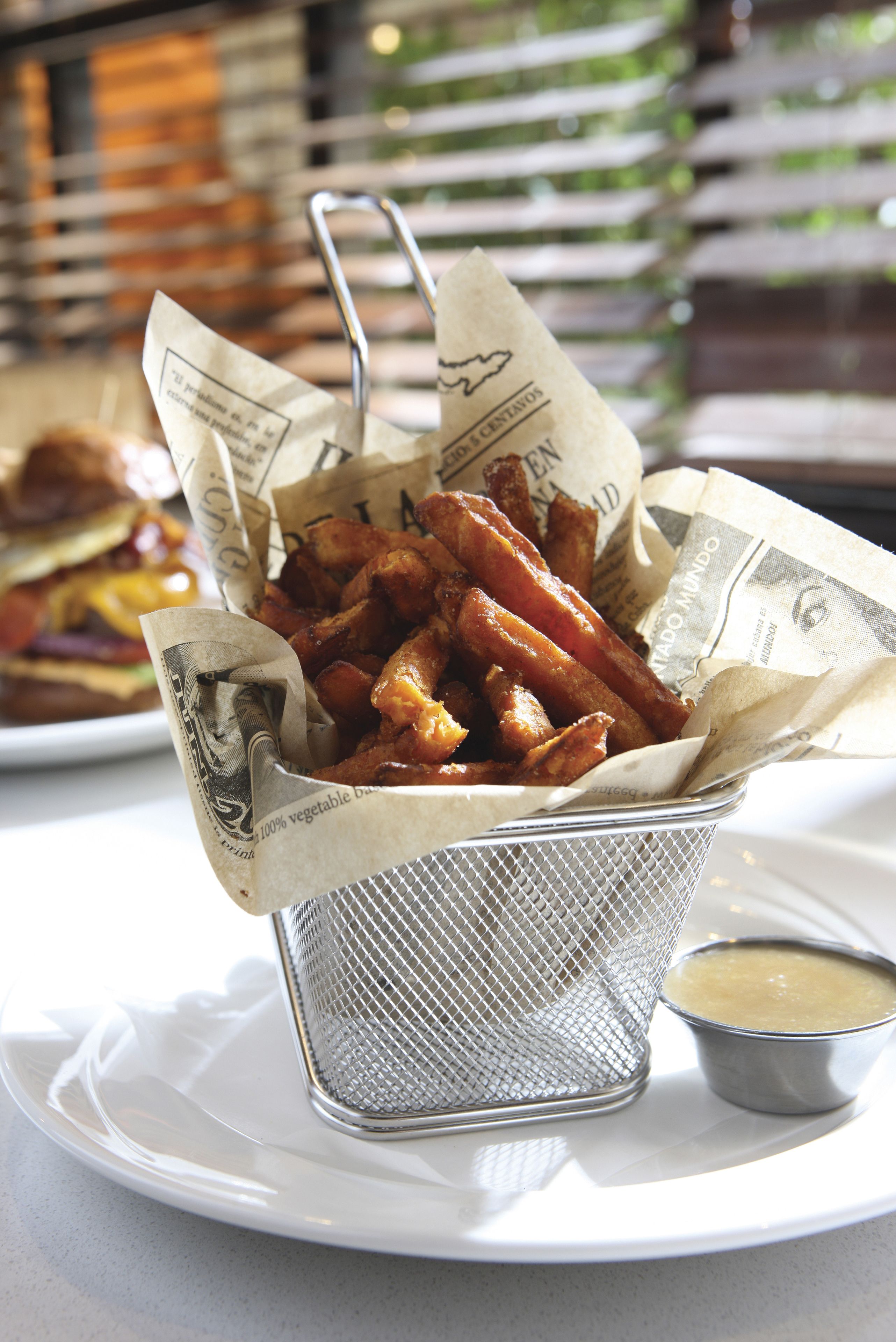 Uma cesta de batatas fritas em recipiente de malha, forrada com jornal, sobre um prato branco.