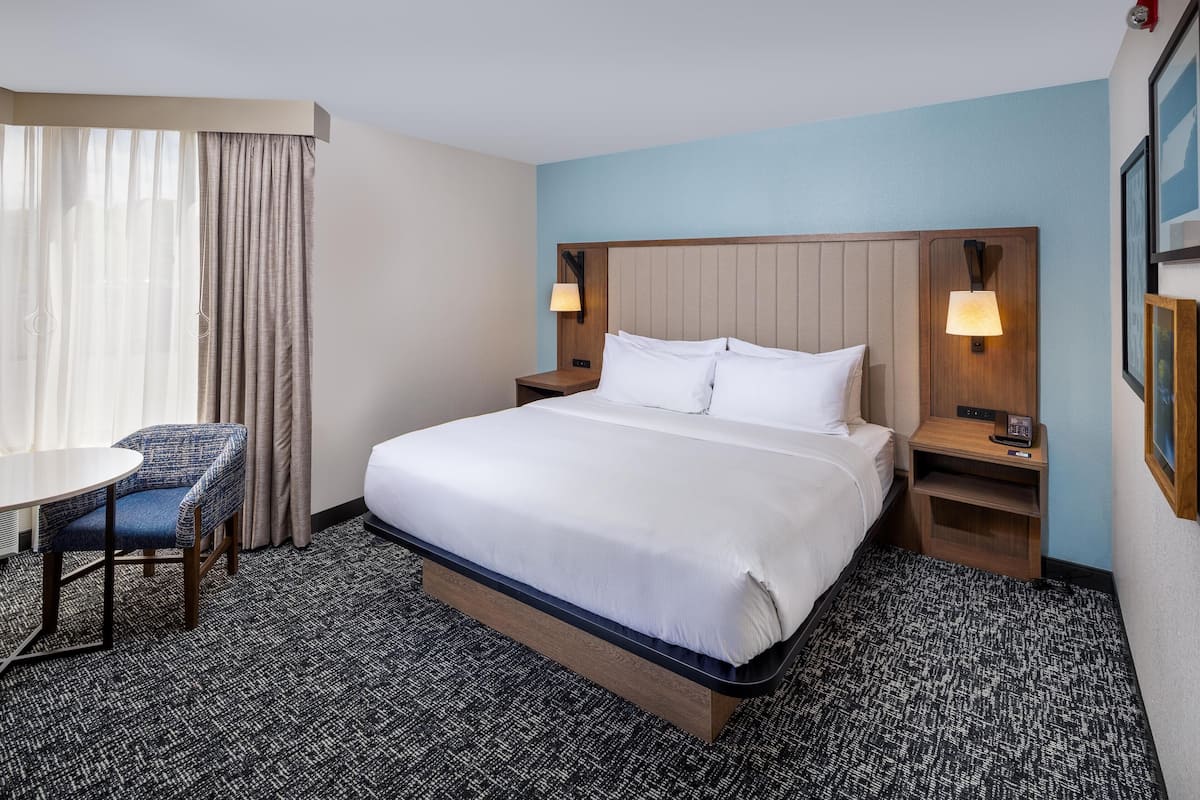 A hotel room with a bed, two bedside tables with lamps, a small table with a chair, and a window with curtains.