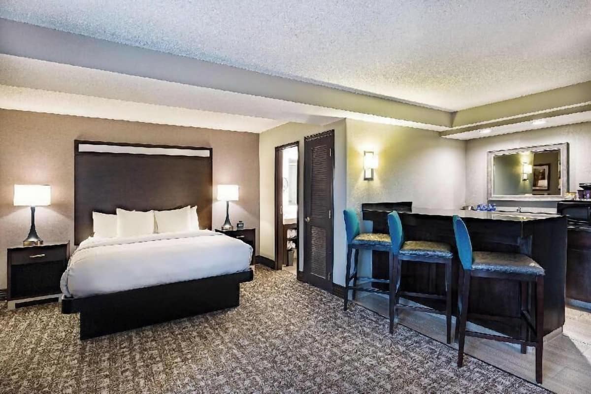 A hotel room with a bed, two bedside tables with lamps, a desk with two chairs, a mirror, and a kitchenette.