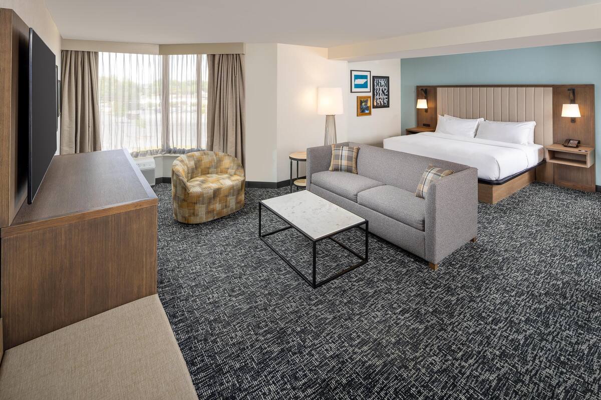 A hotel room with a bed, a sofa, a chair, a TV, a lamp, and a small table.